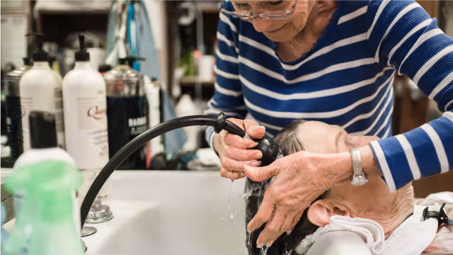 An elderly person is having their hair washed by a caregiver at a sink in what appears to be a salon or care facility. The caregiver is wearing glasses and a blue and white striped shirt, carefully rinsing the person's hair with a handheld sprayer.
