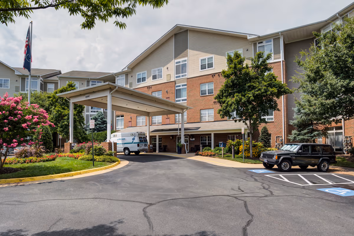 Exterior view of Arbor Terrace Herndon senior living facility showing a multi-story building with a covered entrance driveway. There are trees, flowering bushes, and landscaped greenery around the entrance. A black SUV is parked in a handicapped parking space and an ambulance is parked under the covered entrance.