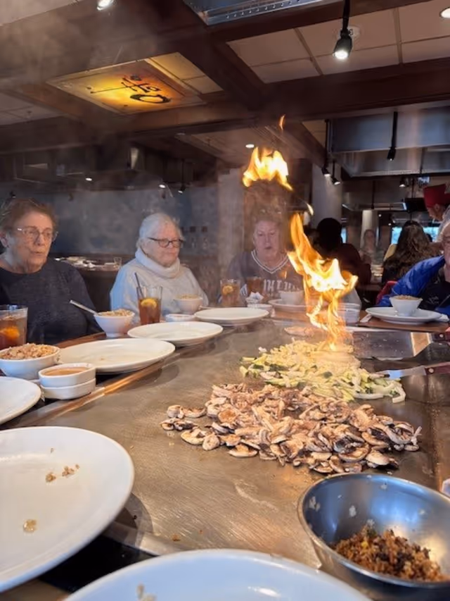 Several elderly people seated around a hibachi grill table in a restaurant, watching a chef cook vegetables and mushrooms with a large flame rising from the grill.