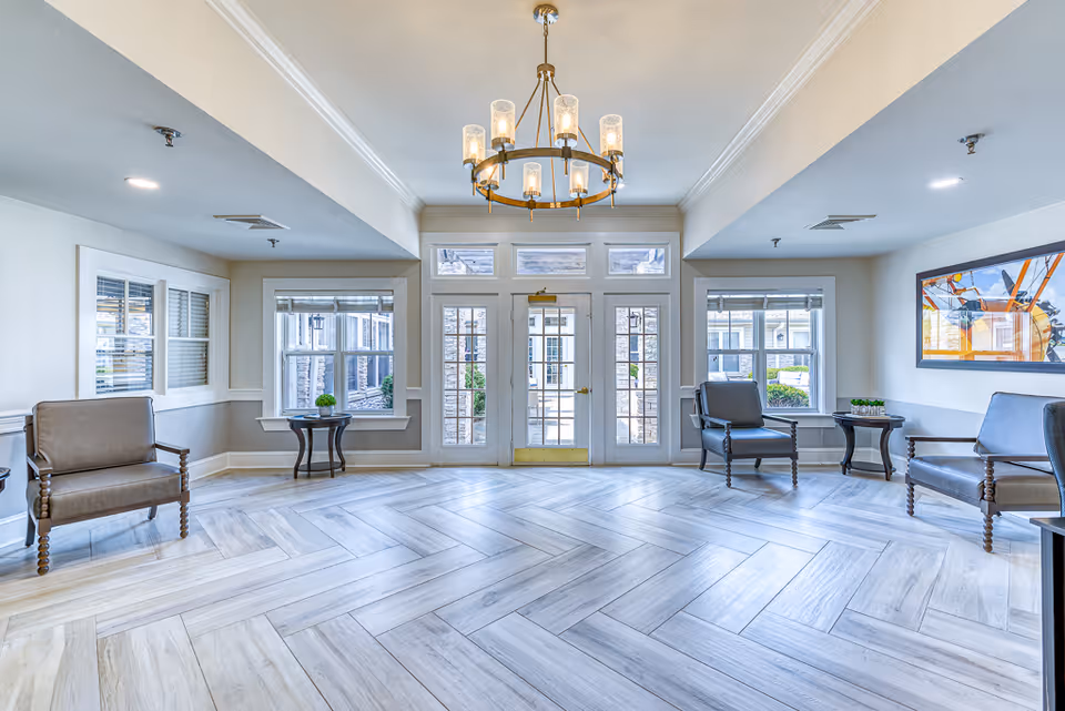 Bright senior living lobby with a central chandelier, seating chairs and side tables, and glass double doors with windows leading outside.