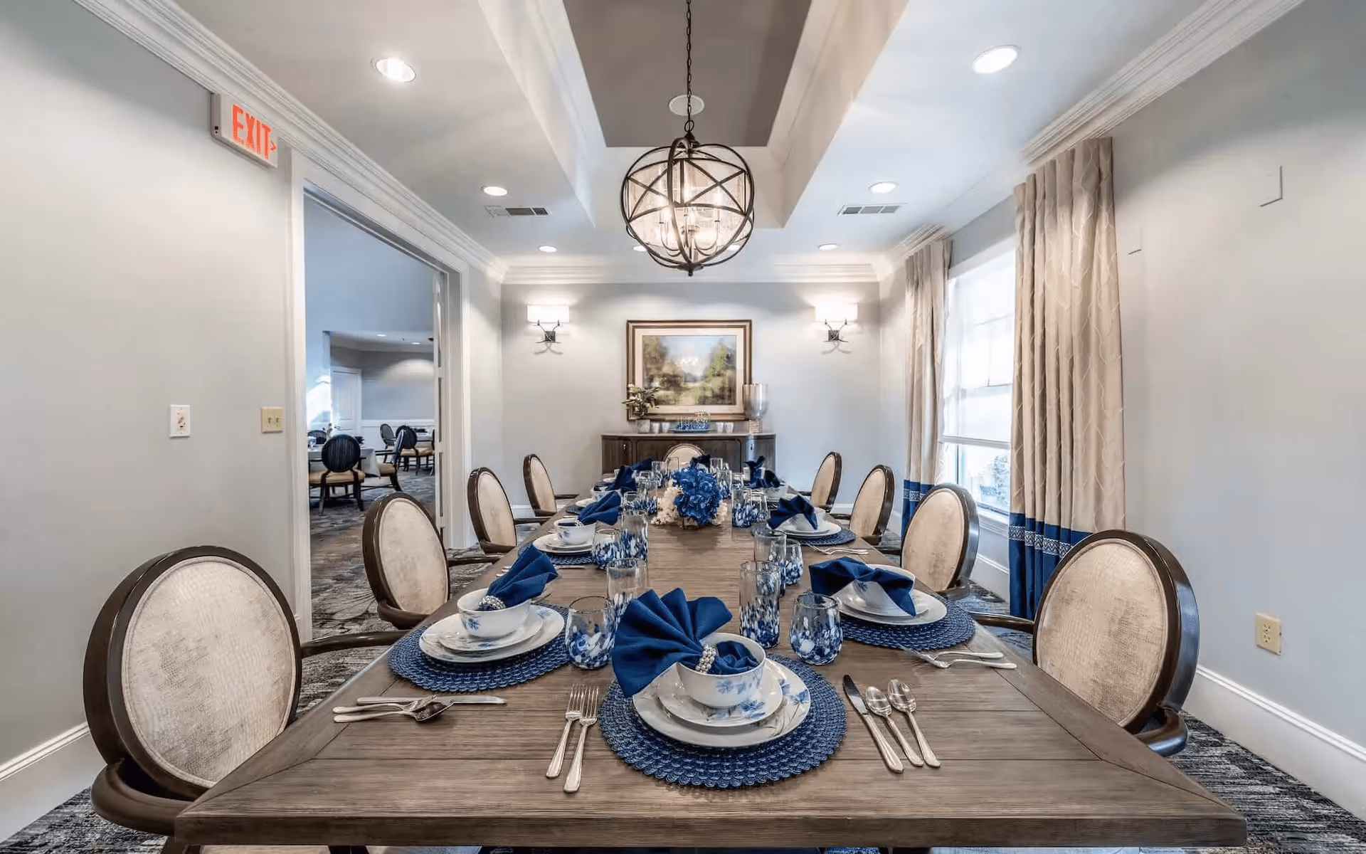 A formal dining room with a long wooden table set for eight people. Each place setting includes a blue woven placemat, white plates with blue patterns, a bowl, silverware, a glass, and a blue folded napkin. The room has beige upholstered chairs with dark wooden frames, light gray walls, a large window with beige curtains featuring a blue band near the bottom, a decorative chandelier hanging from a tray ceiling, wall sconces, and a framed landscape painting on the far wall.