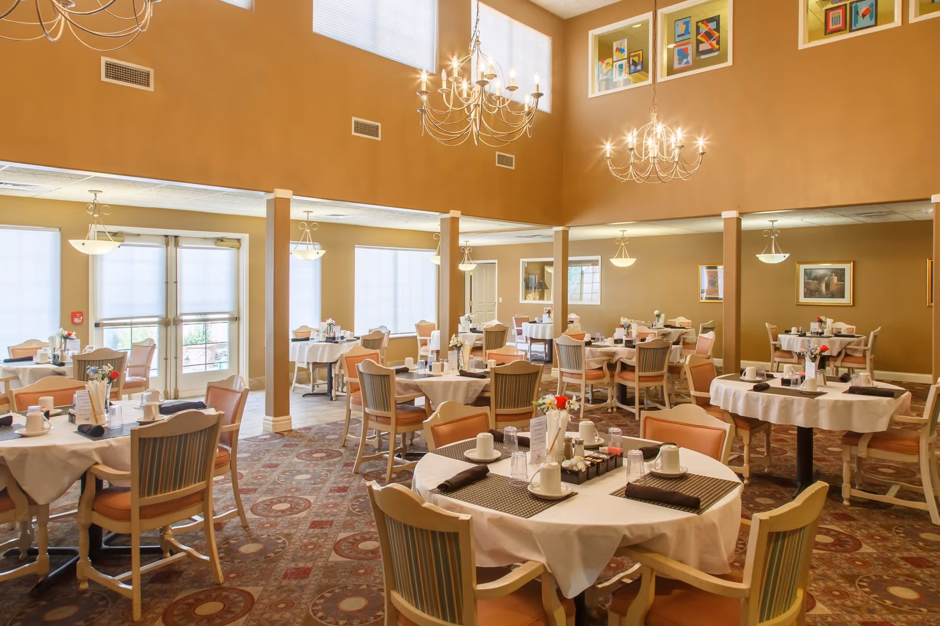 A spacious dining room with multiple round tables covered with white tablecloths, each set with cups, glasses, napkins, and small flower arrangements. The room features high ceilings with two chandeliers, large windows with blinds, and framed artwork on the walls. The carpet has a patterned design, and the chairs have cushioned seats and backs.