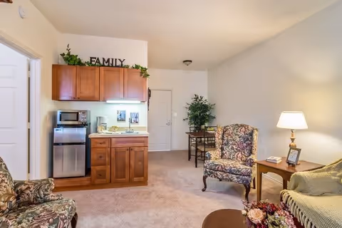 A cozy living area in a senior living facility featuring floral upholstered armchairs, a small wooden side table with a lamp and framed photo, a kitchenette with wooden cabinets, a microwave, and a mini refrigerator. There is a small dining table with chairs near the back wall and decorative greenery on top of the cabinets with the word FAMILY displayed.