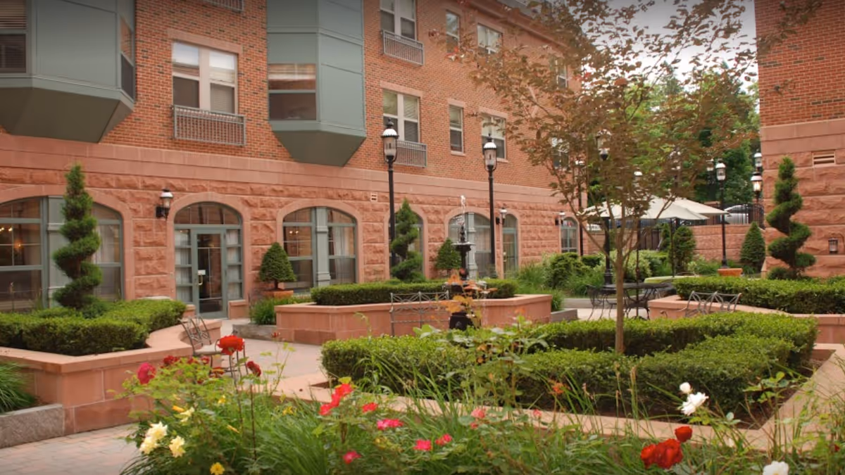 Outdoor courtyard area with neatly trimmed bushes, flowering plants, and small trees. There are benches and tables with umbrellas for seating. The courtyard is surrounded by a brick building with large windows and decorative topiary plants.