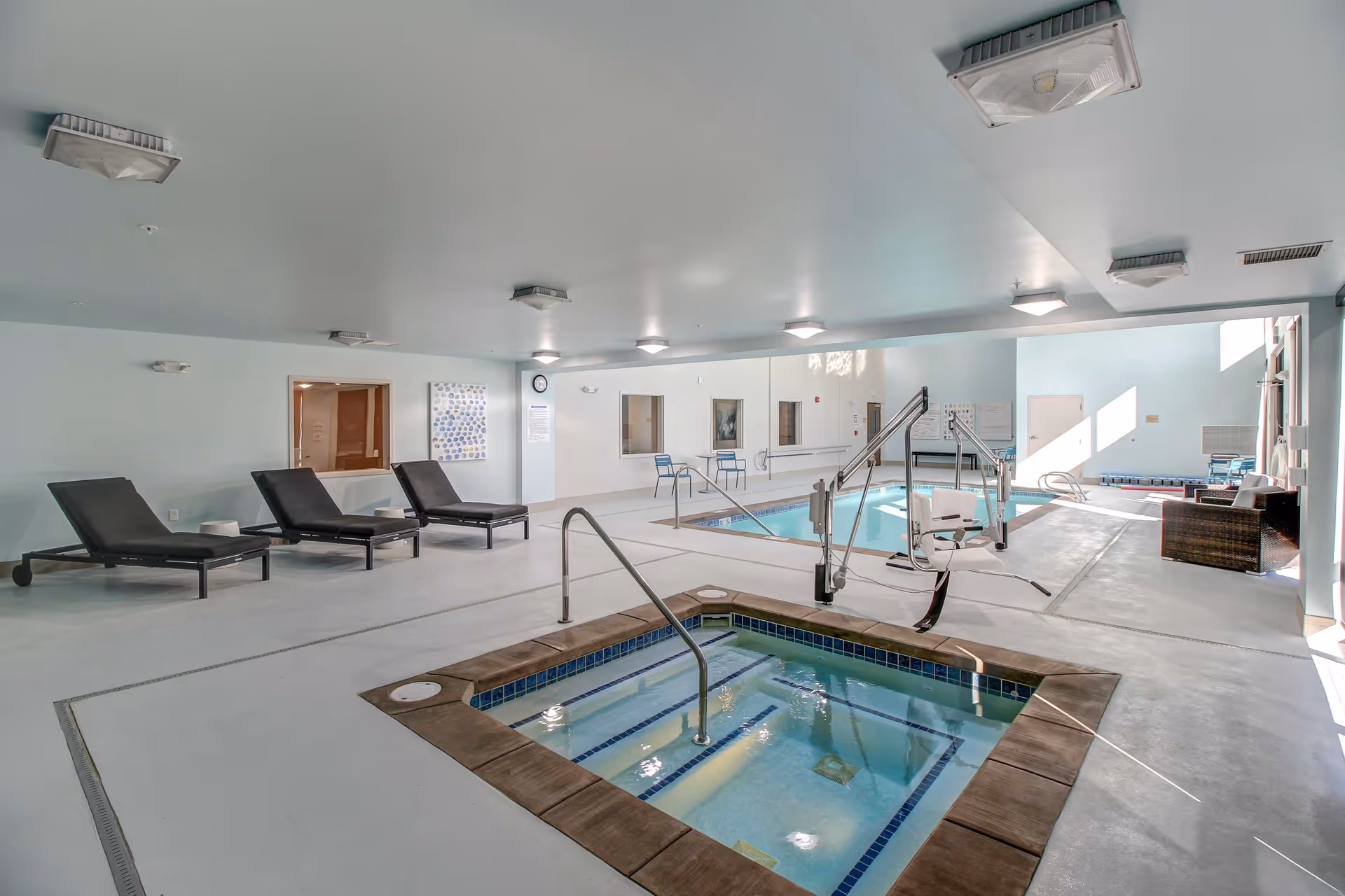 Indoor pool area with a small hot tub in the foreground and a larger swimming pool in the background. There are black lounge chairs along the left wall, metal handrails for pool access, and a pool lift for accessibility. The room has light blue walls and ceiling with multiple ceiling lights, windows, and some chairs near the far wall.