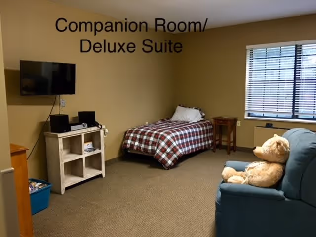 A companion room or deluxe suite in a senior living facility featuring a single bed with a checkered blanket, a small wooden side table, a mounted flat-screen TV above a white shelf unit with electronic devices, a blue armchair with a large teddy bear sitting on it, and a window with blinds letting in natural light.