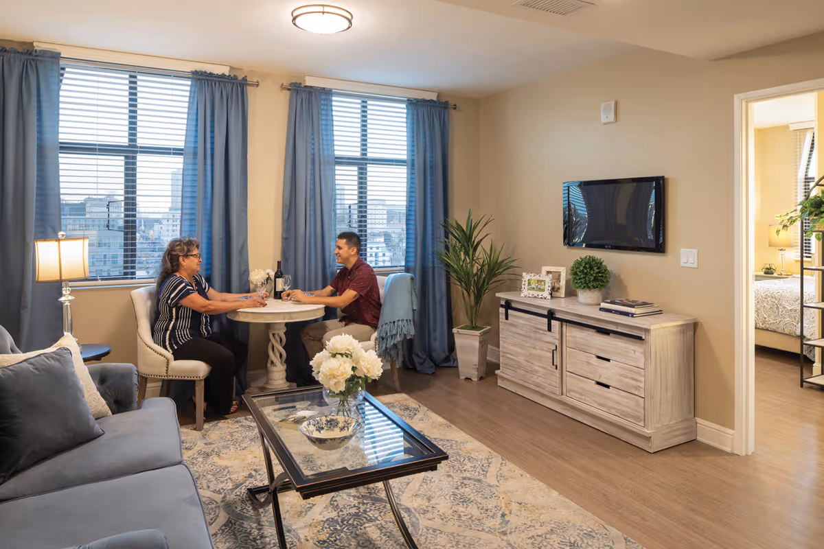 A cozy living room with two people sitting at a small round table near large windows with blue curtains. The room features a gray sofa, a glass coffee table with a flower vase, a wooden cabinet with a mounted flat-screen TV, and a doorway leading to a bedroom.