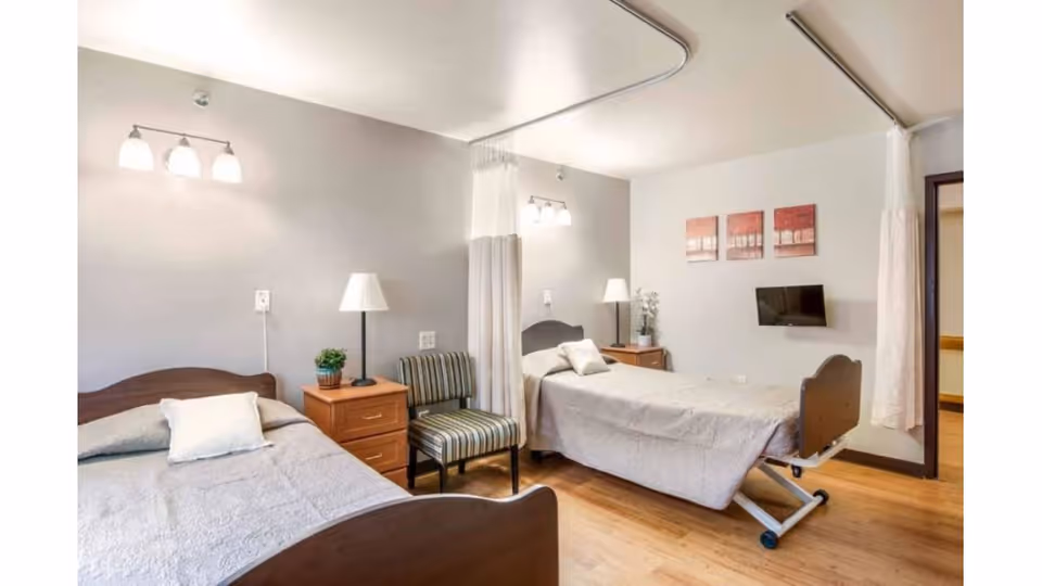 A bright and clean room with two beds, one standard bed and one adjustable hospital-style bed, separated by a privacy curtain. Each bed has a nightstand with a lamp and a small plant. There is a striped chair between the beds, three wall-mounted lights above the beds, and three red and white paintings on the wall. A small flat-screen TV is mounted on the wall opposite the beds. The floor is wooden.