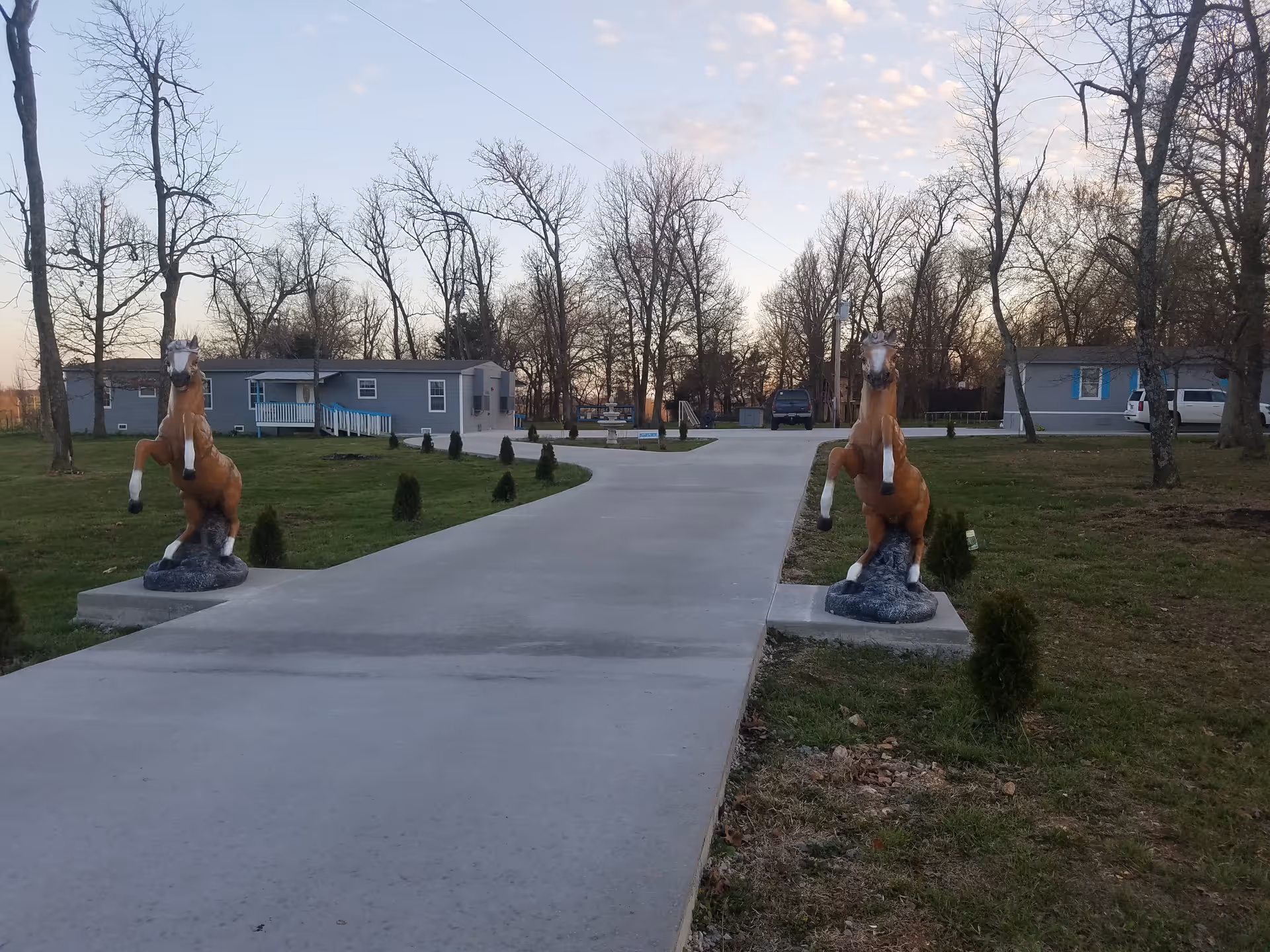 A paved driveway flanked by two rearing horse statues on concrete pedestals, leading to single-story buildings surrounded by grass and leafless trees under a partly cloudy sky.