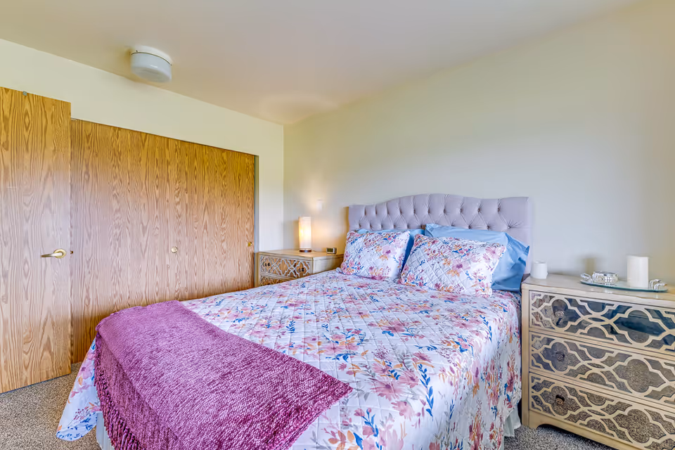 A cozy bedroom with a floral bedspread and purple throw, a tufted headboard, matching bedside tables, and large wooden closet doors.