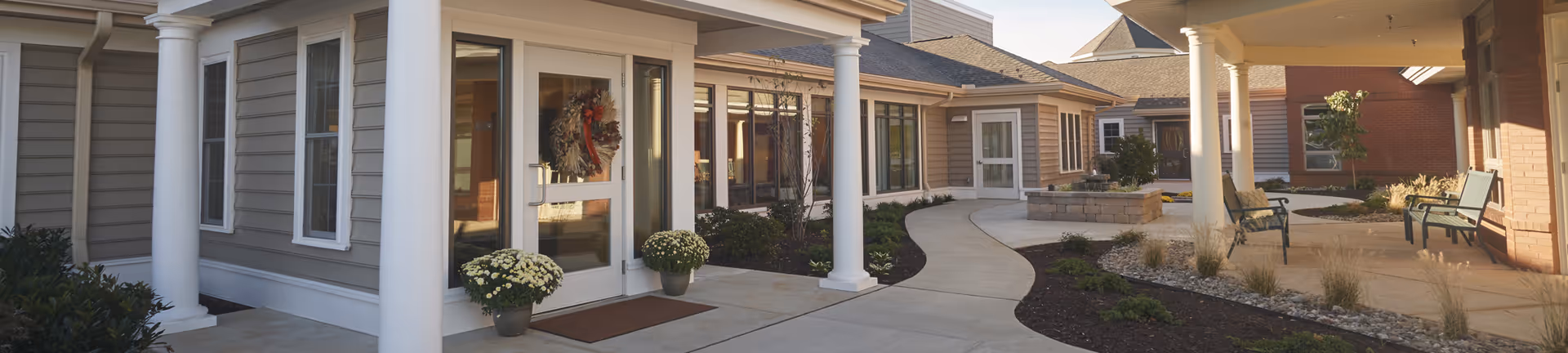 Outdoor view of a senior living facility entrance with a curved concrete walkway, white columns, potted flowers, a decorative wreath on the glass door, and seating areas with benches under a covered patio.