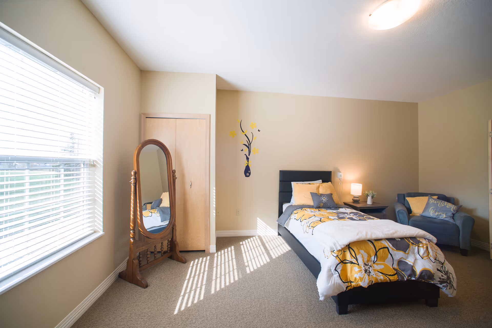 A bright bedroom with a single bed featuring yellow and gray floral bedding, a wooden framed standing mirror, a small wooden nightstand with a lamp and a plant, a blue armchair with a yellow pillow, and a window with blinds letting in sunlight.