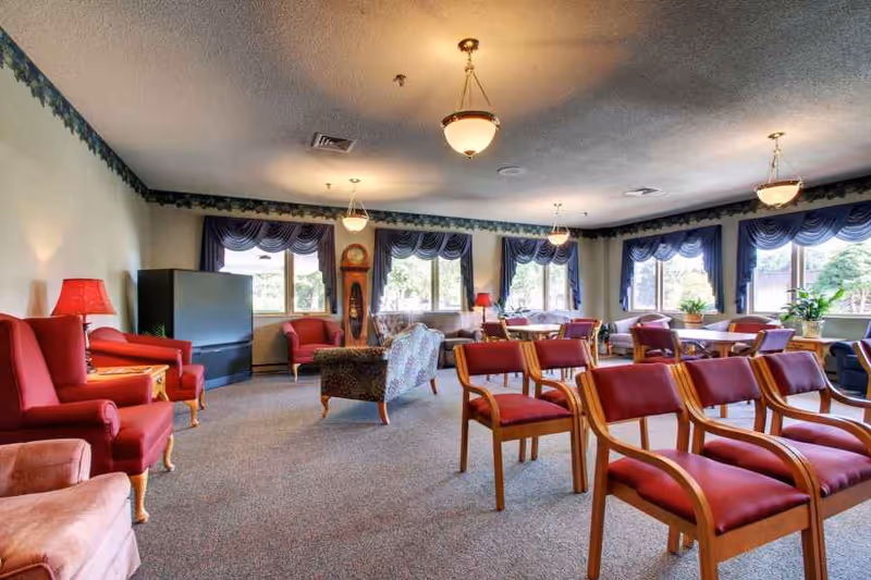 Spacious communal lounge with rows of wooden-framed red chairs, upholstered armchairs, tables, large windows with blue valances, and warm ceiling lights.