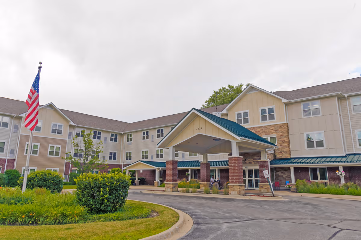 Front entrance of a senior living building with a covered porte-cochère, American flagpole, and landscaped driveway.