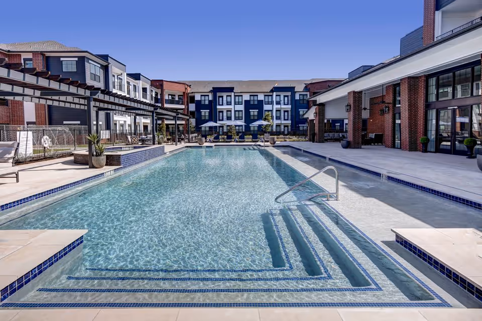 Outdoor swimming pool and poolside patio with seating, surrounded by multi-story residential buildings.