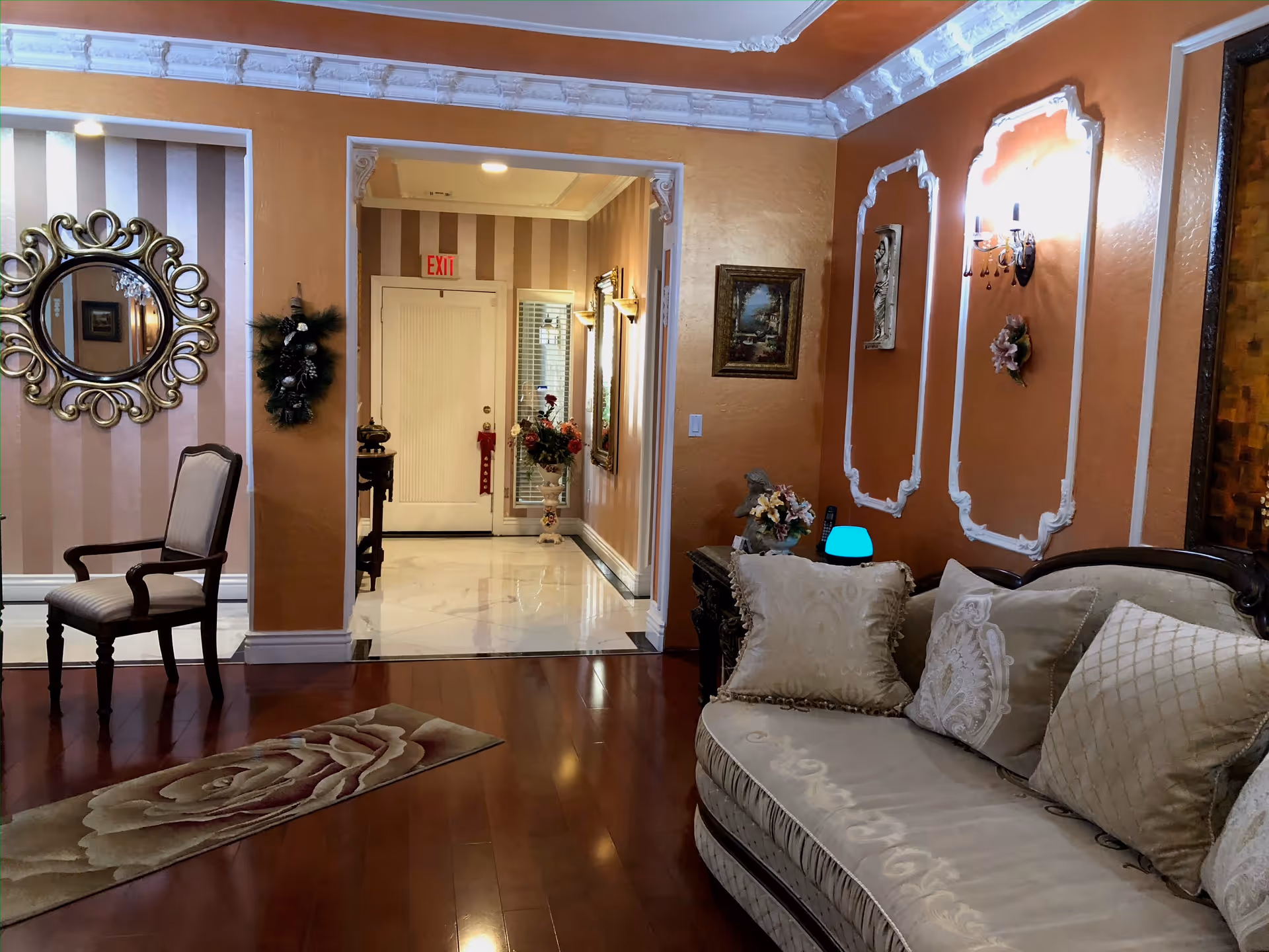 Interior view of a living room area in an assisted living facility with a beige ornate sofa adorned with decorative pillows, a wooden armchair, a floral rug on polished wooden floors, and walls painted in warm tones with white decorative molding. The room opens into a hallway with striped wallpaper, a white door with an exit sign above it, and floral decorations.