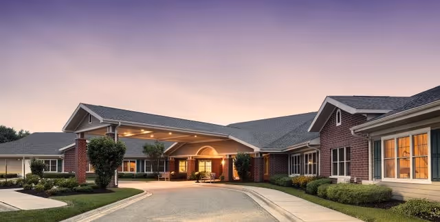 Exterior view of Parkside Manor Assisted Living and Memory Care building at dusk, showing a covered entrance with warm lighting, brick and siding facade, landscaped bushes, and a curved driveway.