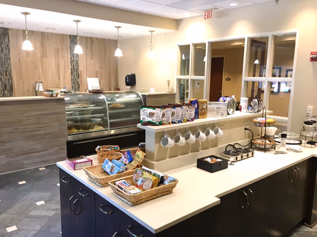Interior view of a snack and beverage counter area in a senior living facility. The counter has baskets filled with various snacks, a row of hanging white mugs, and a display case with baked goods. The background shows a light-colored wall with pendant lights and a hand sanitizer dispenser mounted on the wall.