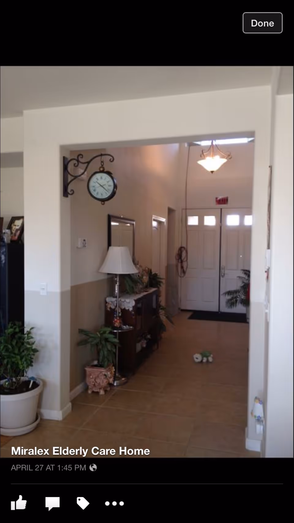 Entry foyer of a care home with tiled floor, a console table and lamp, plants, a hanging wall clock, and front double doors visible at the end of the hallway.