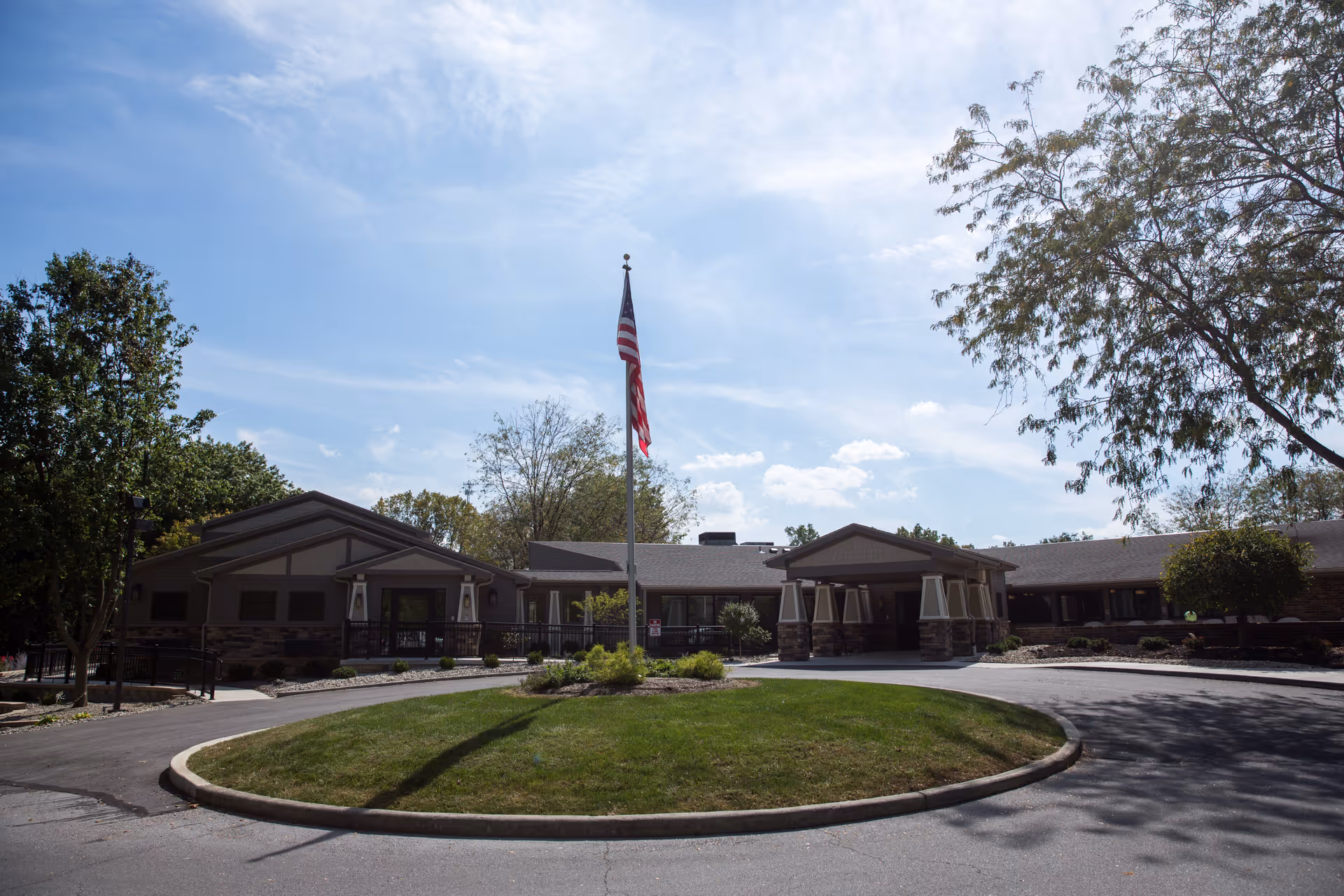 Front entrance of a single-story senior living building with an American flag on a pole, circular driveway and landscaped lawn.