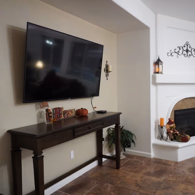 A living room corner featuring a mounted flat-screen TV above a dark wooden console table decorated with autumn-themed items including a small pumpkin and a 'Bless' sign. To the right is a white fireplace mantel adorned with a lantern, a candle, and a small scarecrow figurine. The floor is tiled with brown tiles, and a green plant is placed near the wall.