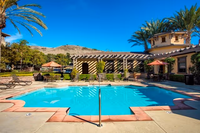 Outdoor swimming pool with lounge chairs, umbrellas, palm trees and surrounding buildings under a clear blue sky.