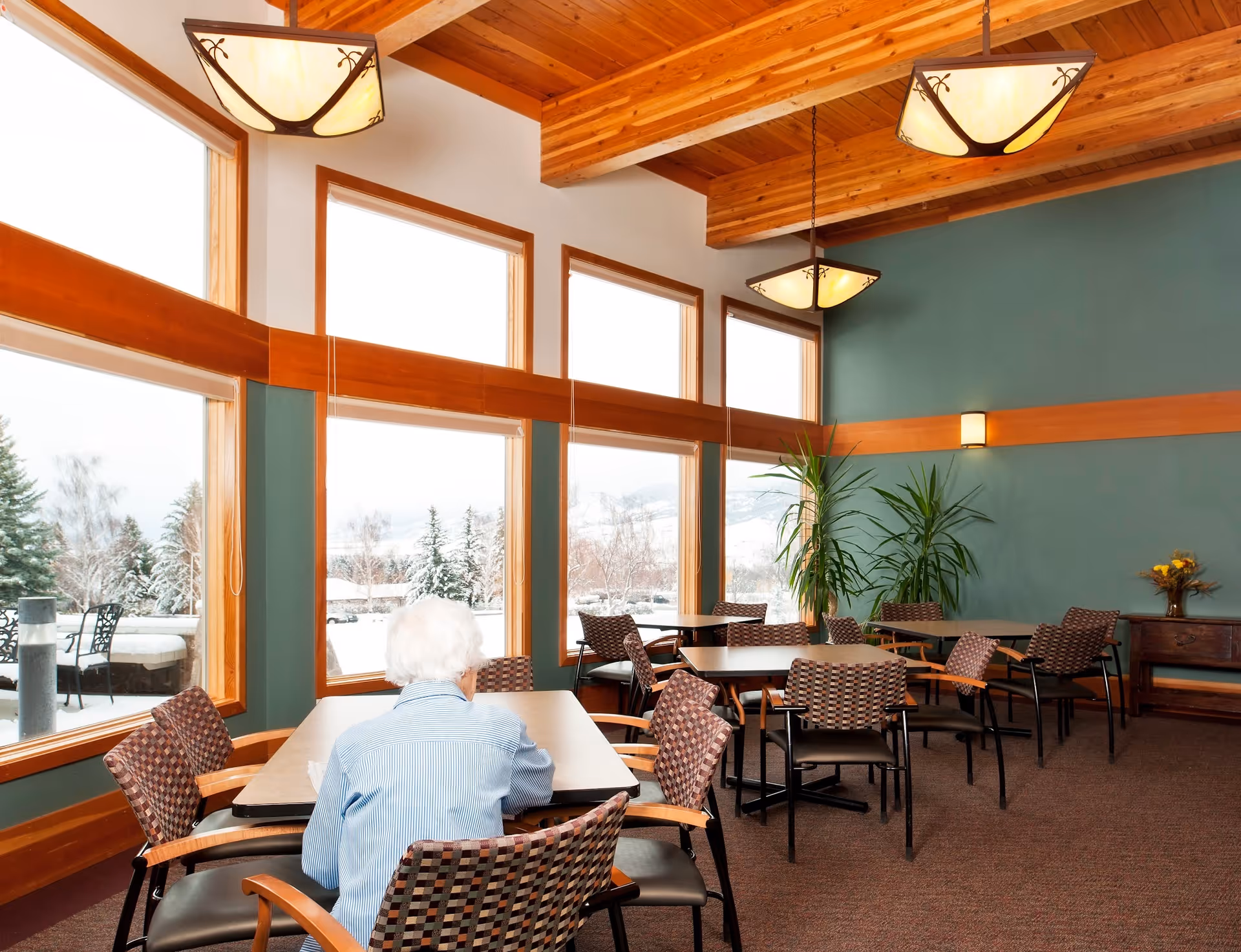 An older adult seated at a table in a bright senior living dining room with large windows looking out to a snowy landscape.