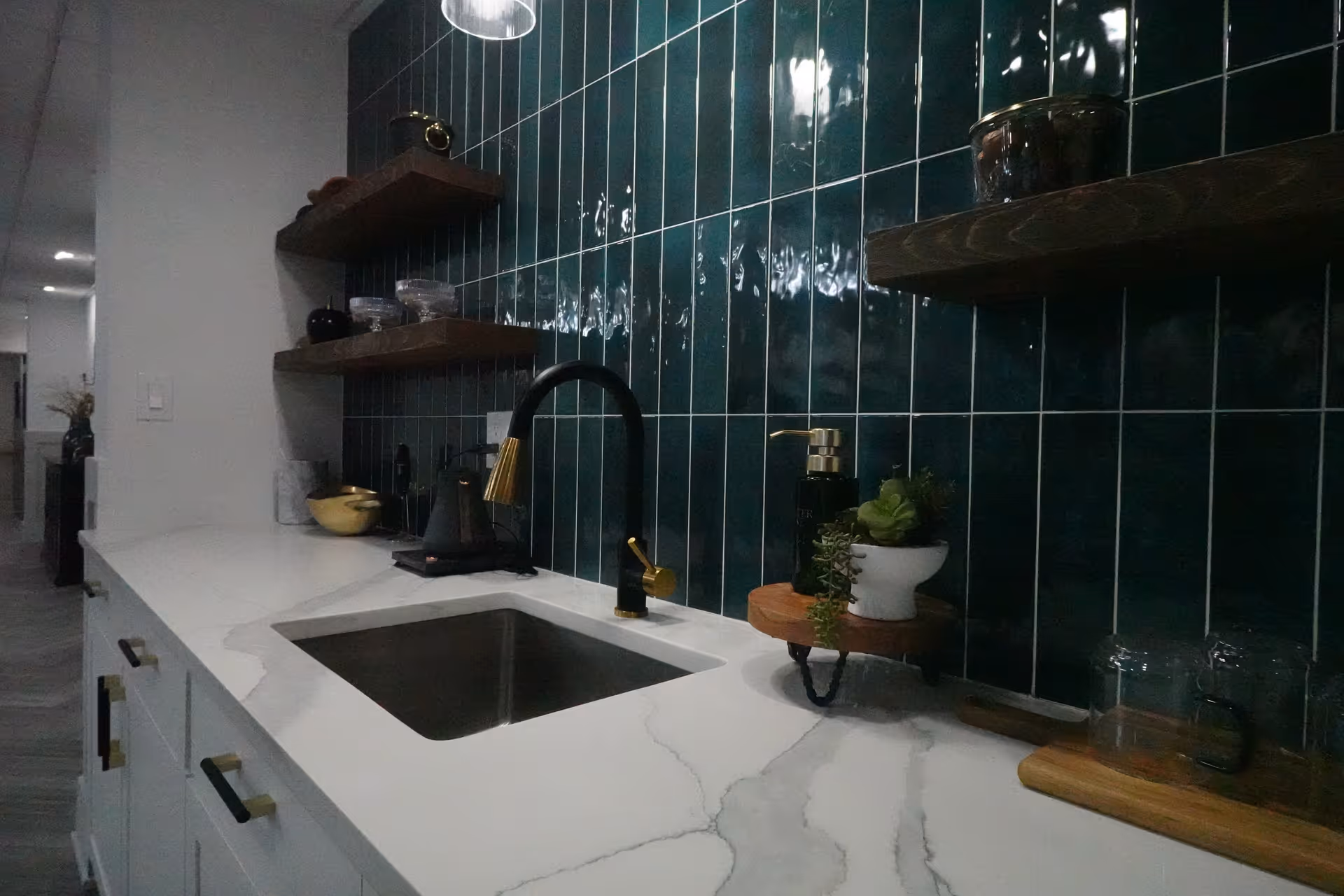 Modern kitchen countertop with a white marble surface, a black and gold faucet over a stainless steel sink, dark green vertical tile backsplash, wooden floating shelves holding small containers and decorative items, and a soap dispenser with a small potted plant on a wooden stand.