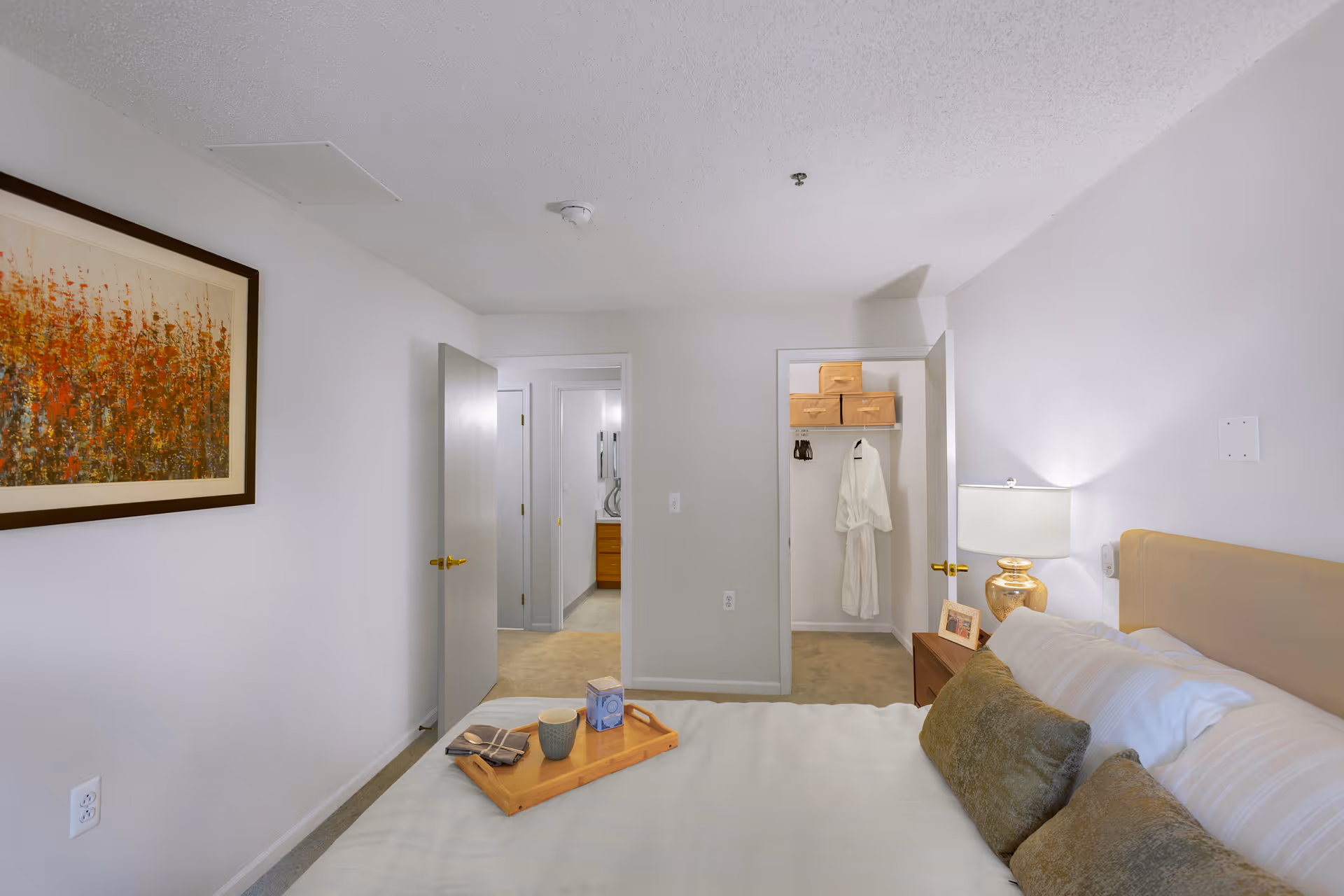 A bedroom in a senior living facility with a neatly made bed featuring white and green pillows. A wooden tray with a cup and a box is placed on the bed. To the right, there is a nightstand with a lamp and a framed photo. The room has light-colored walls, a framed painting on the left wall, and two open doors revealing a closet with storage boxes and a bathrobe, and a bathroom in the background.
