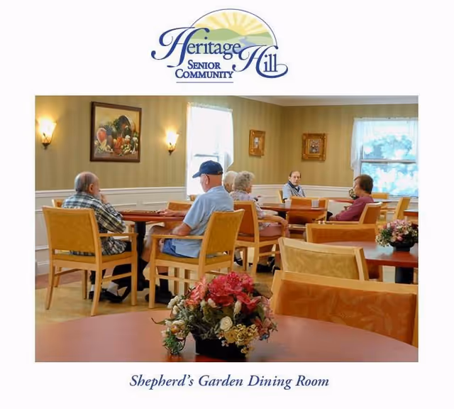 A dining room in Heritage Hill Senior Community with several elderly people seated around tables. The room has beige walls with framed artwork, two windows with white curtains, and floral centerpieces on the tables.