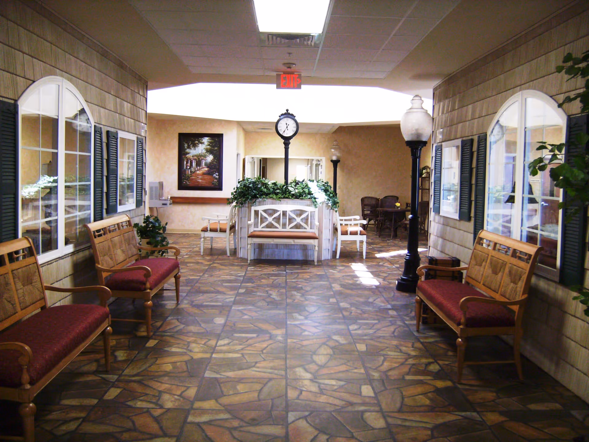 Indoor hallway area with stone-patterned tile floor, wooden benches with red cushions along both sides, decorative street lamps, a clock on a post in the center surrounded by plants, and a seating area with chairs and a table in the background.