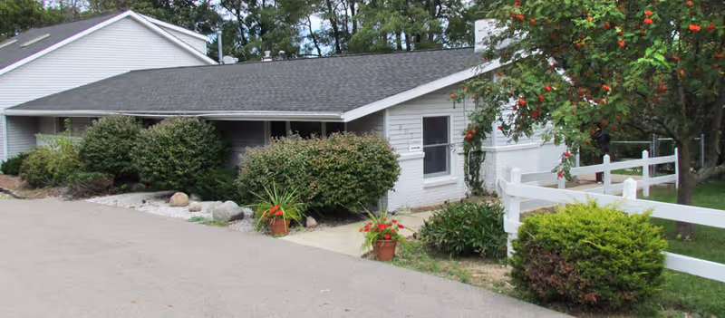 Single-story white brick building with shrubs, potted plants, and a white fence along a paved driveway.