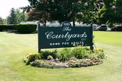 A green entrance sign reading "The Courtyards Home for Adults" sits on a landscaped lawn with trees and grass behind it.