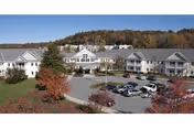 Front exterior of a multi-wing senior living building with a circular driveway, parked cars, and autumn-colored trees.