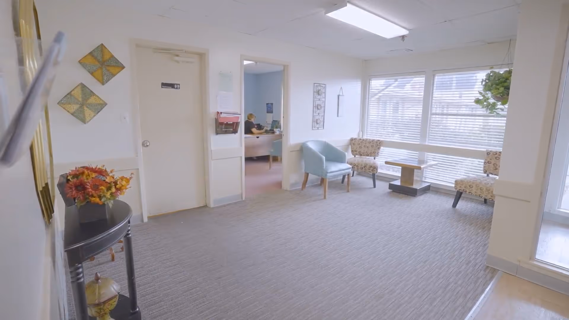 A bright and clean waiting area with a gray carpet, light-colored walls, and large windows with blinds. The room contains a small black table with a flower arrangement, two patterned armchairs, one blue armchair, and a small wooden coffee table. A door labeled 'Restroom' and an open doorway leading to an office with a person working at a desk are visible.