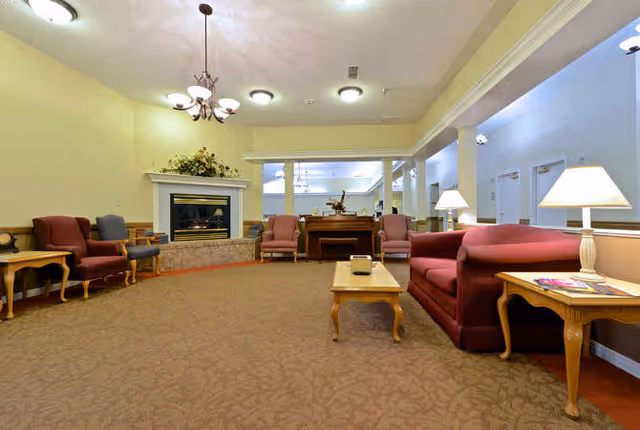 A spacious living room area in Sabine Place featuring a fireplace with floral decoration on the mantel, multiple upholstered armchairs and sofas in red and beige tones, wooden side tables with lamps, a coffee table, and a piano in the background. The room has beige walls, carpeted flooring, and ceiling lights.