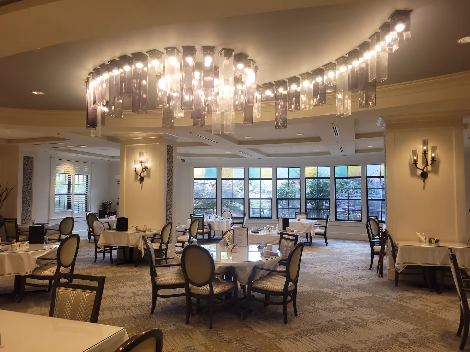 A spacious dining room with round and square tables covered with white tablecloths, surrounded by cushioned chairs. Large windows with stained glass panels allow natural light to enter, and a modern chandelier with multiple hanging lights is mounted on the ceiling. Wall sconces provide additional lighting, and the carpet has a subtle pattern.