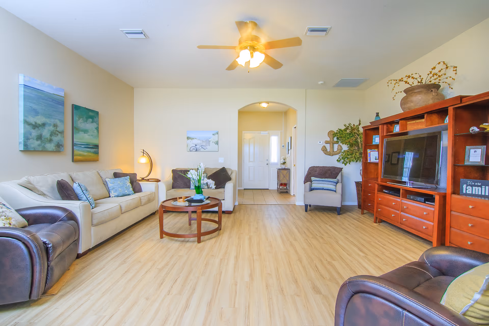 A bright and spacious living room with light wood flooring, a ceiling fan with lights, a large wooden entertainment center with a TV, a beige sofa with blue and brown pillows, a matching loveseat, a dark brown leather armchair, a round wooden coffee table with a flower arrangement, and wall art depicting nature scenes.