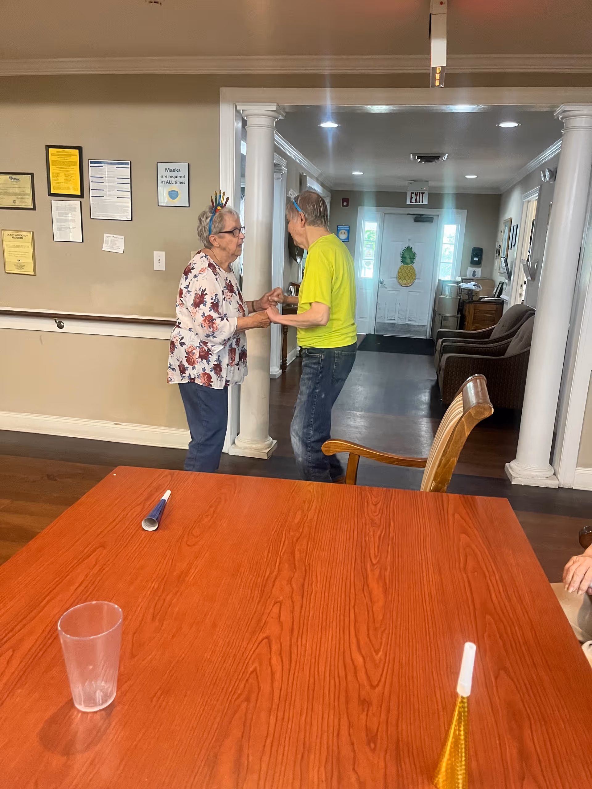 Two elderly people holding hands and facing each other in a hallway of a senior living facility. The hallway has white columns and a door with a pineapple decoration. In the foreground, there is a wooden table with a clear plastic cup and party blowers on it. The walls have several framed notices and a sign that says masks are required at all times.