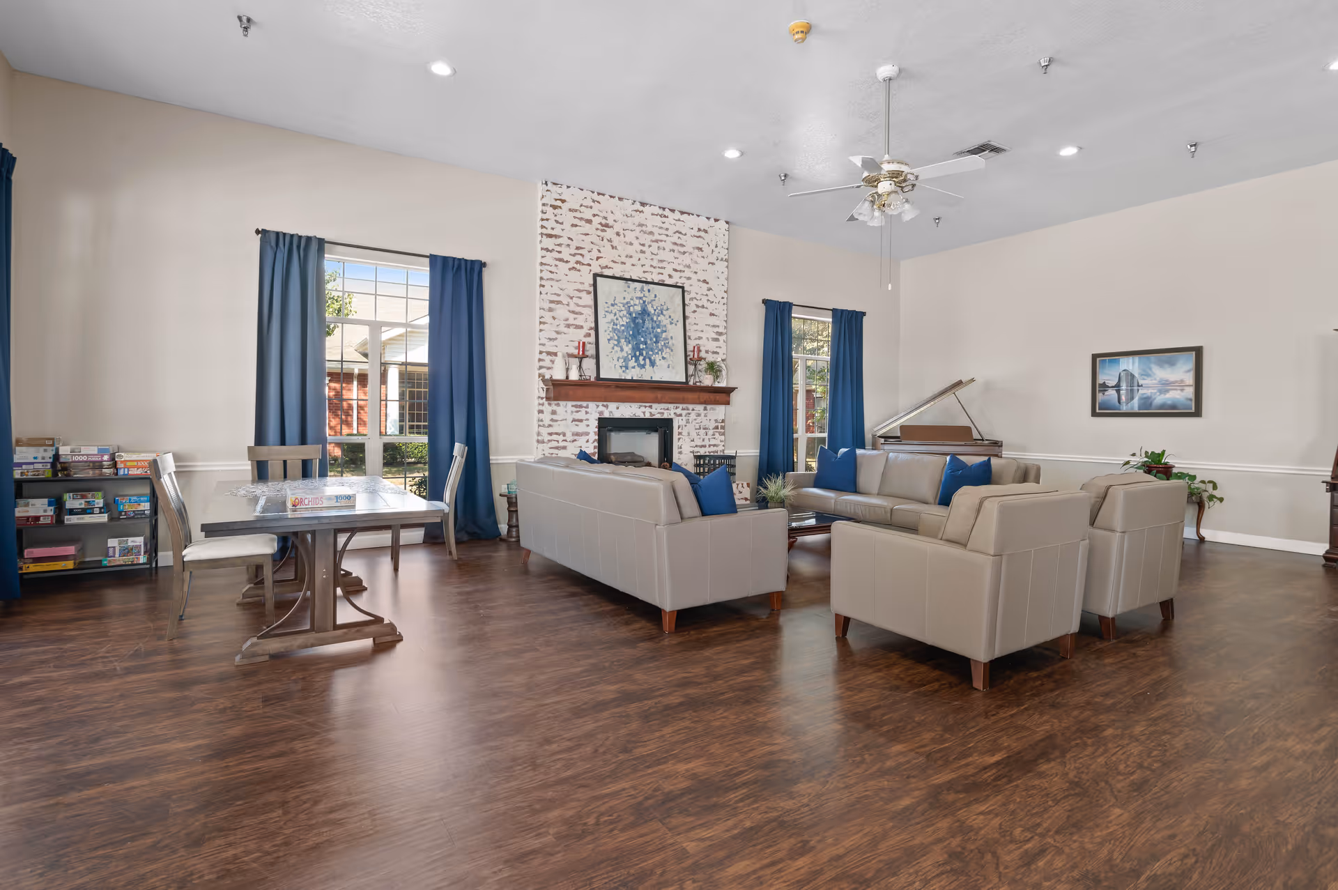 A spacious living room area with beige leather sofas arranged around a coffee table, a white brick fireplace with a painting above it, large windows with blue curtains, a ceiling fan, a piano in the corner, and a table with chairs and board games on shelves nearby.