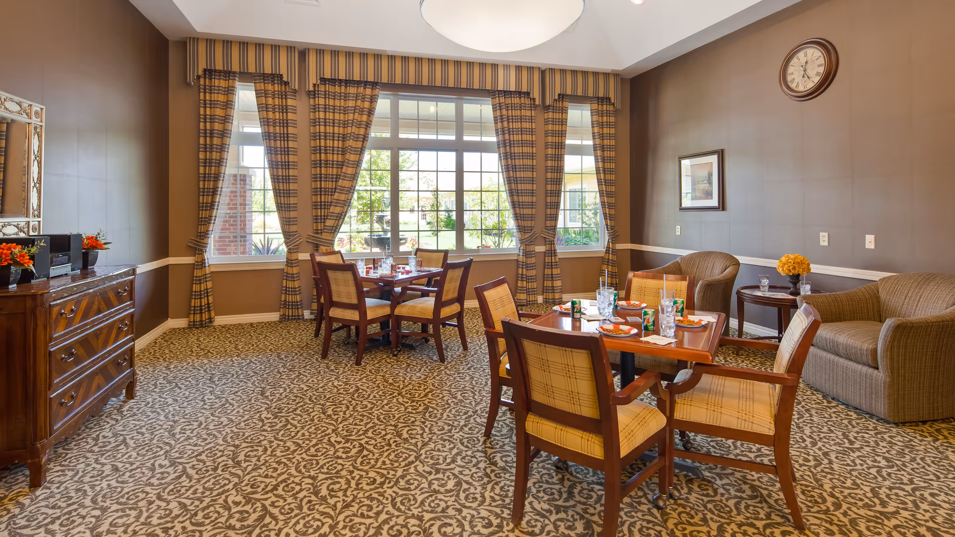 A cozy dining area in a senior living facility with two wooden tables set for meals, surrounded by cushioned chairs with plaid upholstery. The room features large windows with plaid curtains allowing natural light to enter, patterned carpet flooring, a wooden sideboard with decorative items, and a seating area with a sofa and armchairs. A wall clock and framed artwork are visible on the walls.