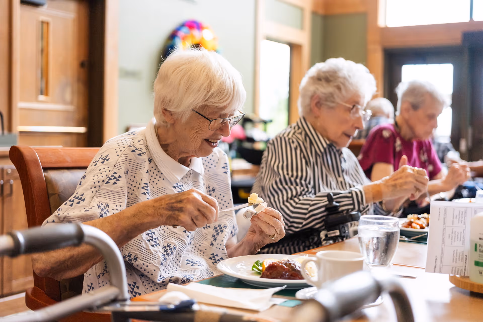 Three elderly women sitting at a dining table in a well-lit room, eating a meal together. The woman in the foreground is smiling and holding a spoon with food. There are plates, glasses of water, and a cup on the table.