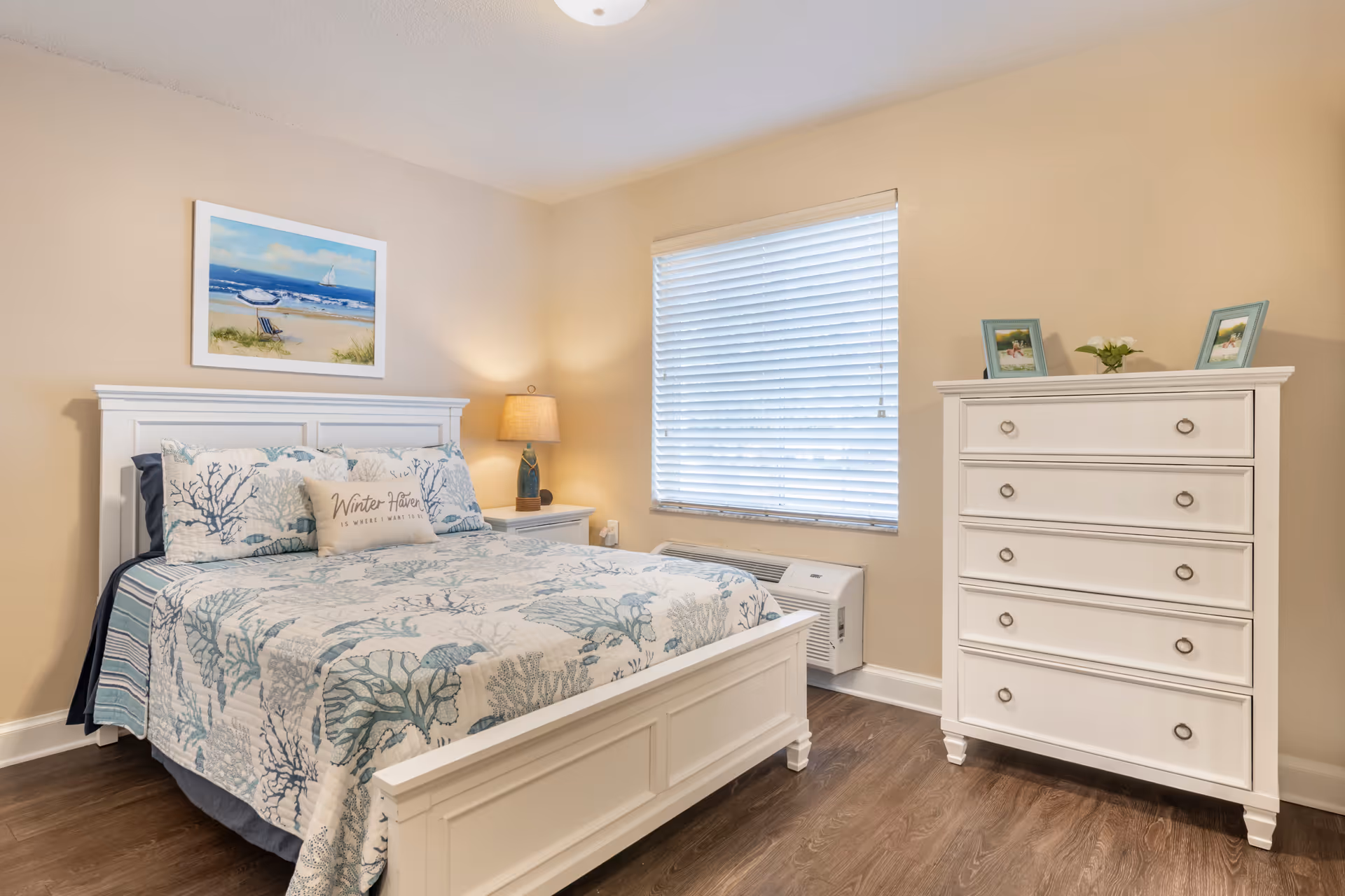 A cozy bedroom with a white wooden bed frame and matching dresser. The bed is made with a blue and white coral-patterned quilt and pillows, including one that says 'Winter Haven is where I want to be.' A nightstand with a lamp and a window with closed blinds are visible. A beach-themed painting hangs above the bed.