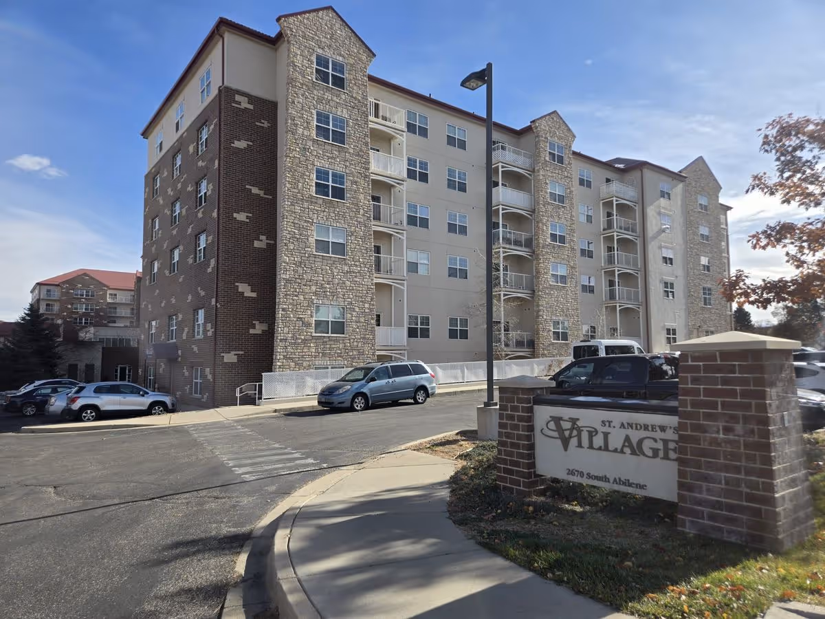 Exterior front view of a multi-story senior living building with stone and brick facades, a driveway with parked cars, and a sign for the village at the entrance.