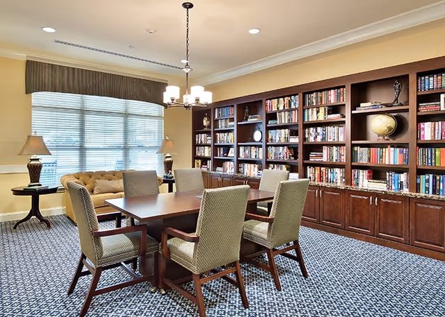 A well-lit library or reading room with a large wooden table surrounded by six upholstered chairs. The room features a wall of built-in wooden bookshelves filled with books and decorative items, a window with blinds and a valance, a beige tufted sofa, two table lamps on side tables, and a patterned carpet.