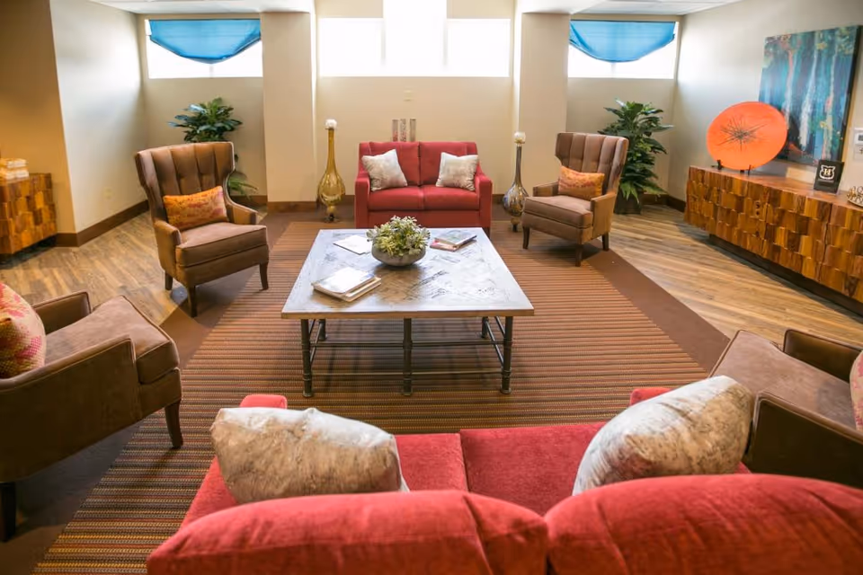 A cozy living room with a red sofa and two brown armchairs arranged around a square wooden coffee table on a striped rug. The room has wooden flooring, two large plants in the corners, blue window shades, and a wooden sideboard with decorative items including a large orange disc and a framed picture.