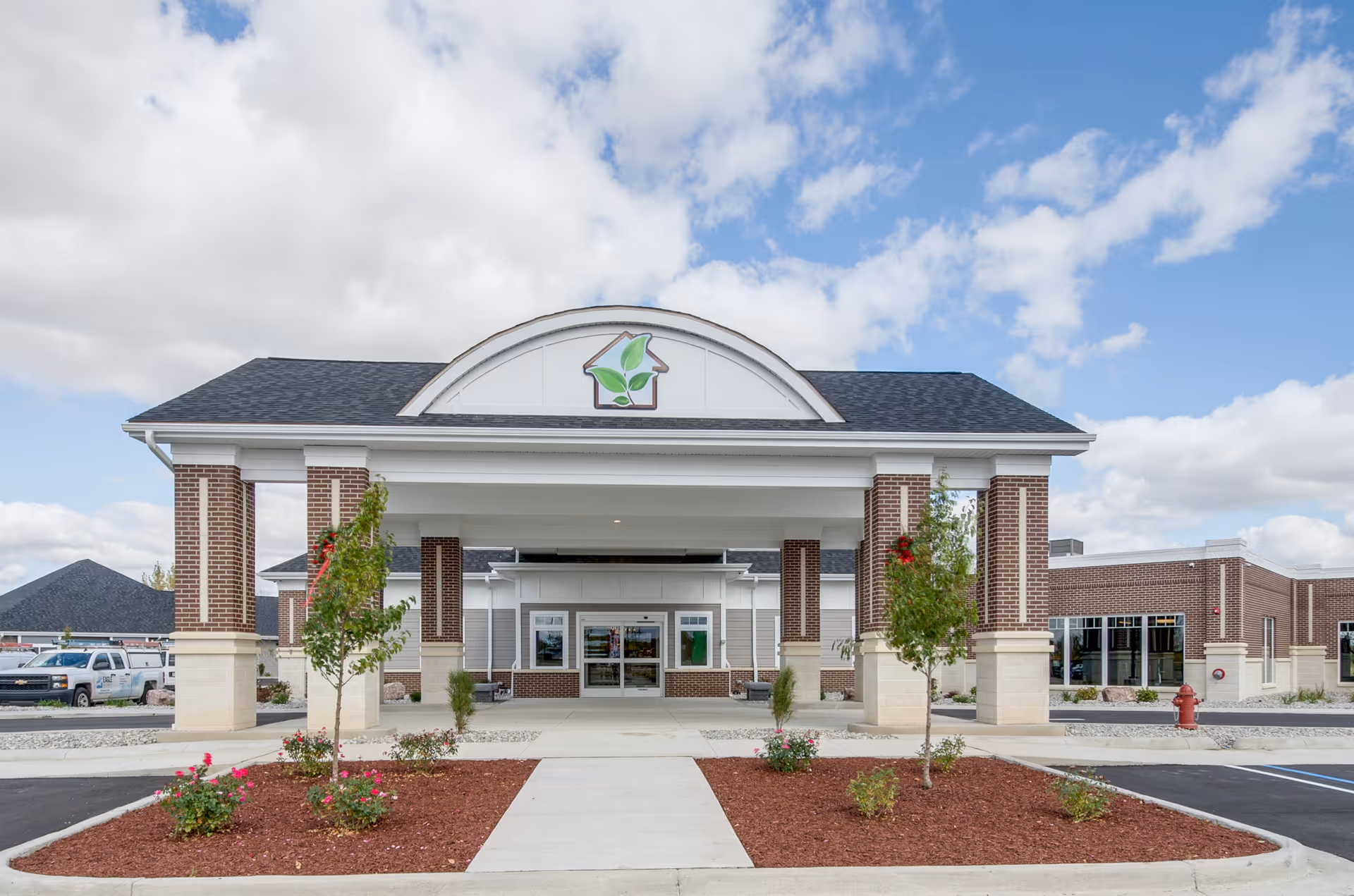 Front exterior view of Great Lakes Rehab Center building with a covered entrance supported by brick and stone columns, landscaped flower beds, and a partly cloudy sky in the background.