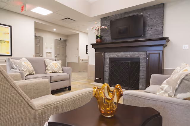 A cozy living room area in an assisted living facility featuring a stone fireplace with a mounted flat-screen TV above it. The room has beige walls and ceiling with recessed lighting. There are two beige armchairs and a beige loveseat with patterned throw pillows arranged around a dark wooden coffee table that holds an amber glass decorative piece. A potted orchid sits on the fireplace mantel. The background shows a hallway with doors and an exit sign.