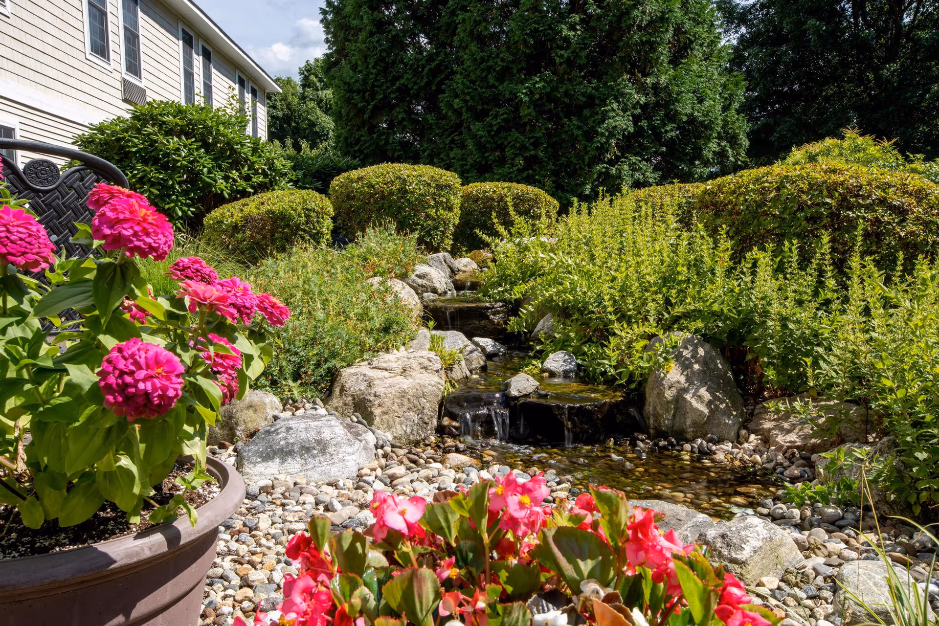 A peaceful garden area at East Village Place featuring vibrant pink flowers in pots, a small flowing water stream surrounded by rocks, neatly trimmed bushes, and lush green trees under a partly cloudy sky.