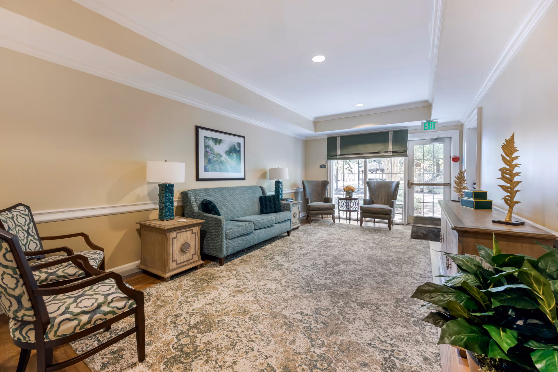 A well-lit living room area with a patterned carpet, a teal sofa with two matching pillows, two wooden side tables each with a blue lamp, two patterned armchairs on the left, and two wingback chairs near a glass door with a green valance. There is a wooden cabinet on the right with decorative golden leaf sculptures and a green plant in the foreground. A framed artwork hangs above the sofa.